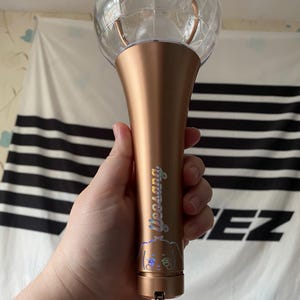 K&ouml;nnte beinhalten: Ein ros&eacute;goldener Lichtstick mit einer durchsichtigen Plastikkugel oben. Der Lichtstick hat einen holografischen Aufkleber mit dem Text "Yeosang" und einer Comicfigur.