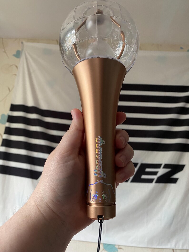 K&ouml;nnte beinhalten: Ein ros&eacute;goldener Lichtstick mit einer durchsichtigen Plastikkugel oben. Der Lichtstick hat einen holografischen Aufkleber mit dem Text "Yeosang" und einer Comicfigur.