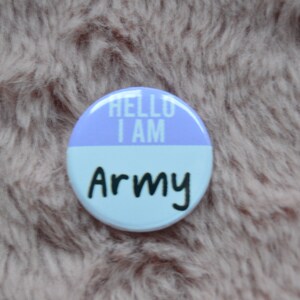 Puede incluir: Un botón redondo con fondo morado y blanco. El botón dice "Hello I am Army" en texto negro.