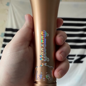 K&ouml;nnte beinhalten: Ein goldener Lichtstick mit einem holografischen Aufkleber eines Cartoon-Hundes und dem Wort "Stray Kids" darauf.