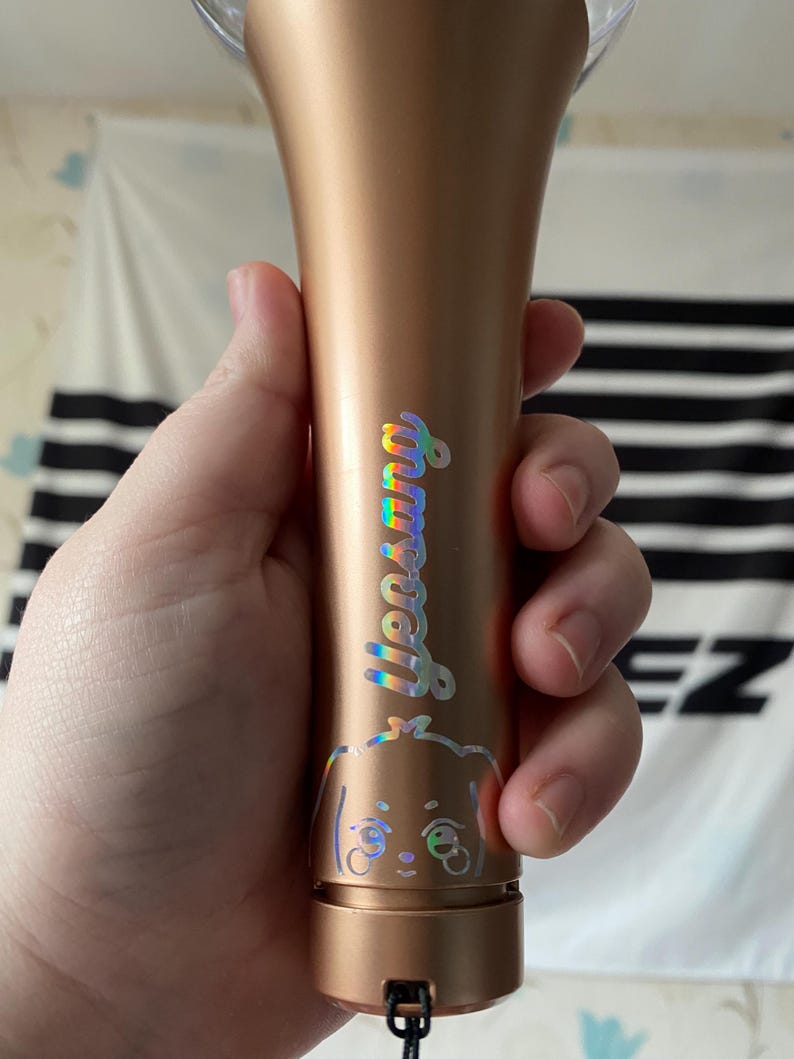 K&ouml;nnte beinhalten: Ein goldener Lichtstick mit einem holografischen Aufkleber eines Cartoon-Hundes und dem Wort "Stray Kids" darauf.