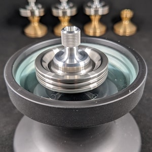 May include: A silver metal spinning top with a black base and a clear glass lid. The top is designed with a rounded, smooth surface and a central shaft.