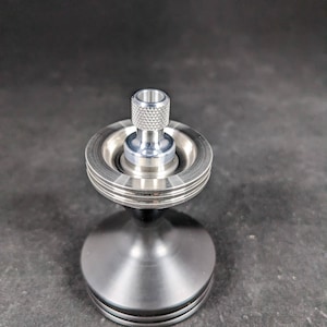 May include: A silver metal spinning top with a textured top and a wide base. The top is designed to spin on a central axis.