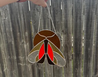 Puede incluir: Atrapasoles de vidriera con forma de polilla, con una combinación de colores marrón, rojo y gris. El diseño de la polilla está suspendido por una cadena plateada. Se muestra frente a una valla de madera desgastada.