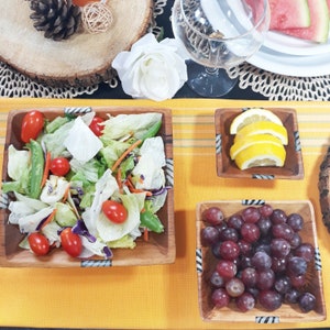 May include: Wooden square bowls filled with fresh salad, sliced lemons, and grapes. The salad includes lettuce, tomatoes, and snap peas. The bowls are on a yellow tablecloth, with a wine glass and watermelon slices in the background.