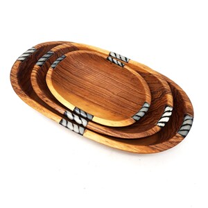 May include: A set of three wooden oval serving bowls. The bowls are nested, with the largest at the bottom and the smallest at the top. The wood grain is visible. Each bowl has decorative bands with a black and white pattern.