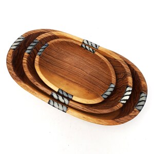May include: A set of three wooden oval serving trays. The trays are nested, with a light brown wood grain finish. Each tray has decorative accents of white and grey stripes on the ends.