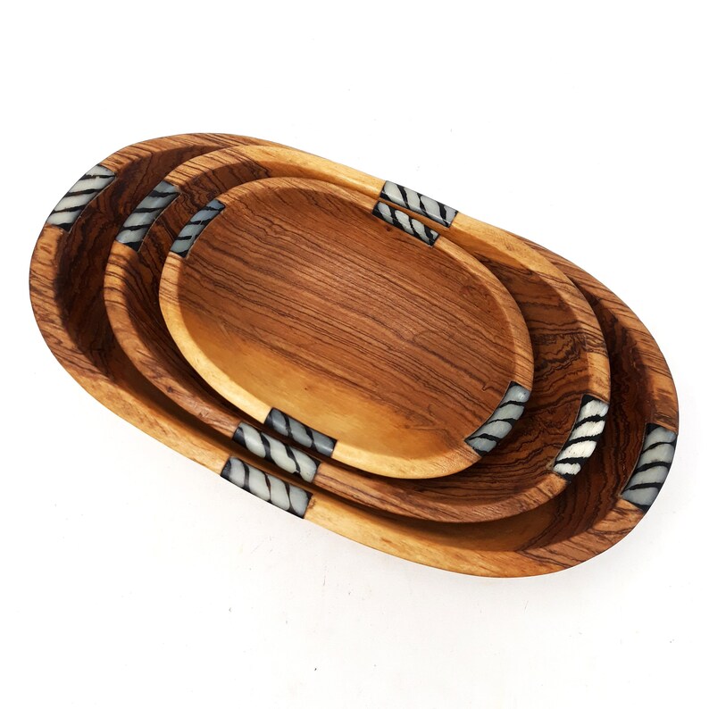 May include: A set of three wooden oval serving trays. The trays are nested, with a light brown wood grain finish. Each tray has decorative accents of white and grey stripes on the ends.