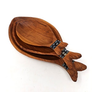 May include: A set of three wooden serving spoons in a fish design. The spoons are made of brown wood and have a decorative band of black and white dots near the handle. The spoons are nested together. The spoons are made of wood.