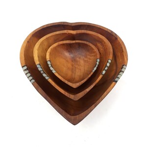 May include: A set of four wooden heart-shaped bowls nested together. The bowls are made of a warm-toned wood and have decorative inlaid accents. The bowls vary in size, with the largest bowl at the bottom and the smallest at the top.