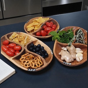 Puede incluir: Tres fuentes de madera para servir con aperitivos. Una fuente contiene totopos y tomates cherry. Otra tiene patatas fritas, tomates cherry, aceitunas negras y pretzels. La tercera contiene hierbas, jengibre y ajo.