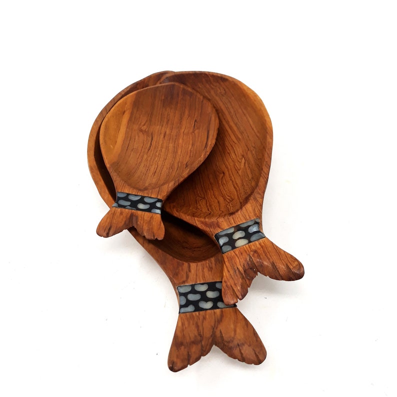 May include: Wooden serving spoons and a serving bowl. The spoons and bowl are made of a warm brown wood. The spoons have a decorative band of black and white stones near the handle. The bowl is round and has a similar wood grain.