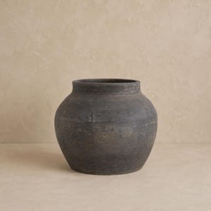 May include: A dark gray, round ceramic pot with a wide opening and a slightly textured surface. The pot has a simple, classic design and appears to be handmade. The neutral color and shape make it suitable for various decor styles.