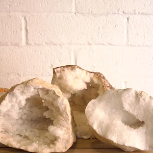 May include: Three geode specimens with a rough, beige exterior and a sparkling, crystal-filled interior. The geodes are displayed on a wooden surface against a white brick wall, showcasing their natural beauty and unique formations.
