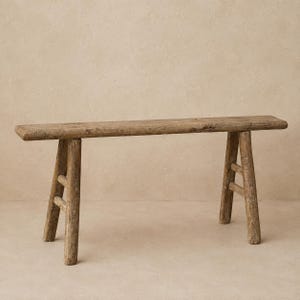 May include: A long, narrow bench constructed from weathered, light brown wood. The bench features a flat, rectangular seat supported by four splayed legs, with two horizontal support bars connecting the legs on each side of the structure.