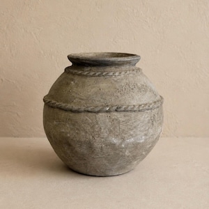 May include: A gray, textured, round vase with a rope-like design around the middle. The vase has a wide, open top and a slightly darker rim. The neutral color and rustic texture give it a natural, earthy aesthetic.