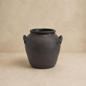May include: A matte black ceramic vessel with a wide, rounded body and a short, thick neck. The pot features two small, curved handles positioned on opposite sides near the rim, showcasing a textured, dark finish with subtle variations in tone.