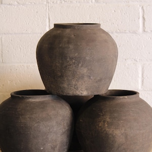 Puede incluir: Tres ollas redondas de cerámica gris oscuro con amplias aberturas están apiladas sobre una superficie de madera contra una pared de ladrillos blancos. Las ollas tienen un aspecto rústico y envejecido, lo que sugiere que son vintage o hechas a mano.