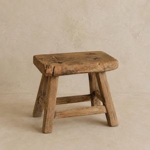 May include: A small, rectangular wooden stool with a weathered, brown finish. The stool features a thick, solid top surface with visible grain and natural imperfections, supported by four sturdy, slightly angled legs connected by horizontal stretchers.