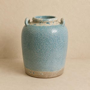 May include: A ceramic vase with a rounded body and two small handles at the neck. The surface features a textured, crackled glaze in shades of light blue and turquoise, transitioning to a smooth, unglazed beige base at the bottom rim.