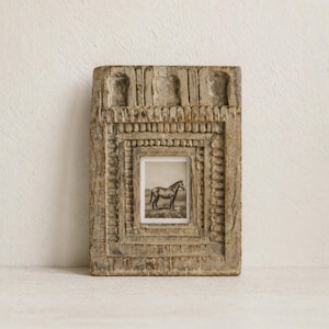 May include: A rectangular, weathered wooden frame with intricate carved details. The frame surrounds a small, black and white image of a horse standing in a field. The frame has a rustic, antique appearance.