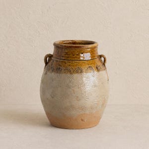 May include: A large, handcrafted ceramic vase with a wide, rounded body and two small handles. The vase features a two-tone color scheme, with a light beige base and a rich brown top. The body is decorated with a repeating floral pattern.