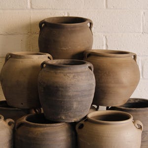 May include: A collection of rustic, handcrafted pottery vessels in varying shades of brown and gray. The pots have a rounded shape with two small handles on either side. They are stacked in a visually appealing arrangement against a white brick wall.