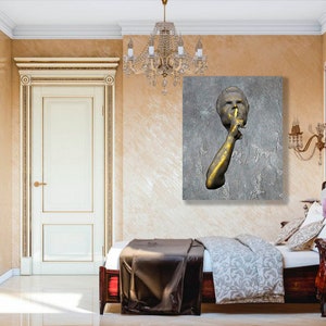 May include: A gold-toned sculpture of a person with their finger to their lips, set against a grey textured background. The sculpture is displayed on a wall in a bedroom with a bed and bedside table.