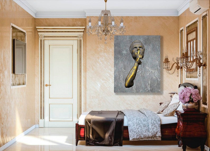 May include: A gold-toned sculpture of a person with their finger to their lips, set against a grey textured background. The sculpture is displayed on a wall in a bedroom with a bed and bedside table.