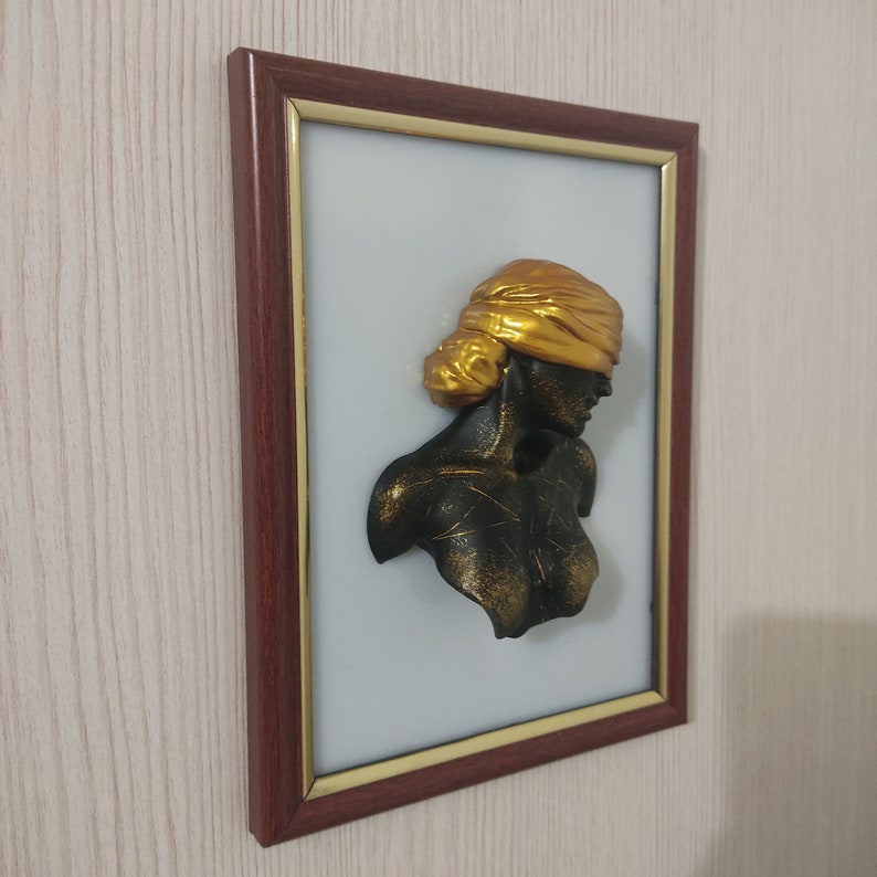 May include: A framed 3D wall sculpture of a woman's bust with a gold blindfold. The sculpture is painted black with gold accents and is set against a white background.