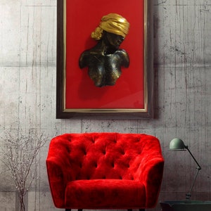 May include: A framed wall sculpture of a woman's bust with a gold head covering. The sculpture is black and has a textured finish. The frame is brown wood and the background is red.