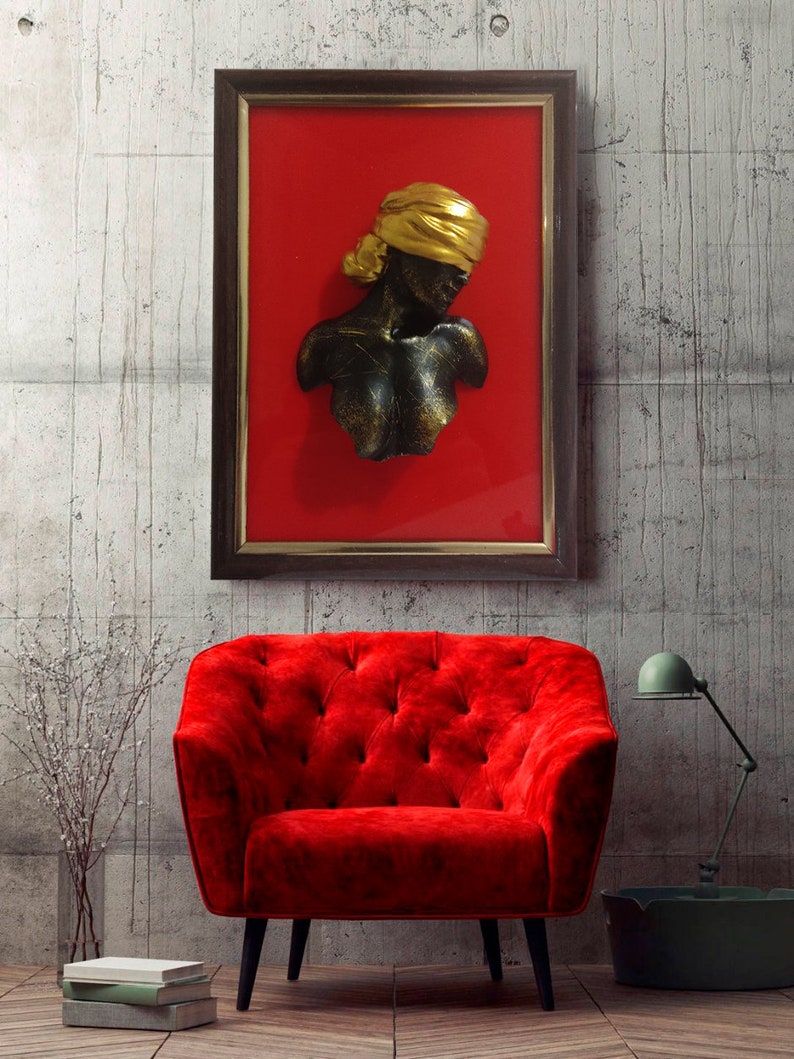 May include: A framed wall sculpture of a woman's bust with a gold head covering. The sculpture is black and has a textured finish. The frame is brown wood and the background is red.