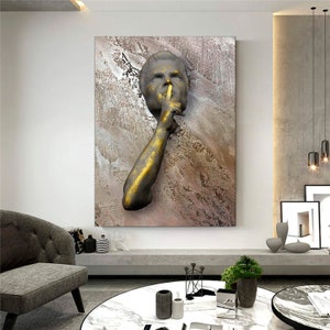 May include: A gold and brown abstract wall art piece featuring a 3D sculpture of a person's face with their finger to their lips, as if to say "shhh".