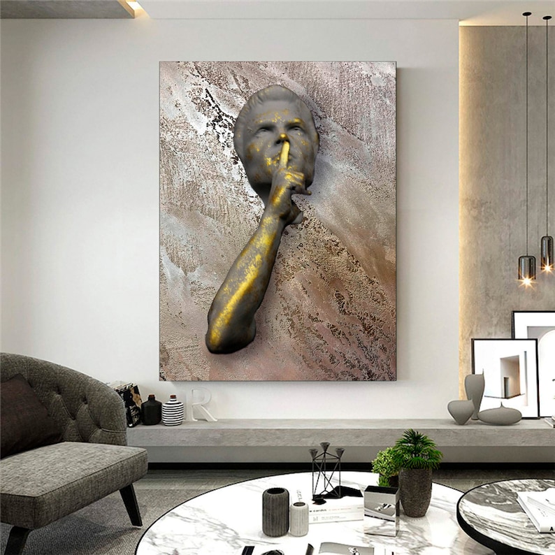 May include: A gold and brown abstract wall art piece featuring a 3D sculpture of a person's face with their finger to their lips, as if to say "shhh".