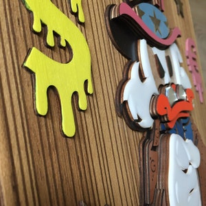 May include: A wooden wall art piece featuring a cartoon character wearing a cowboy hat and glasses. The character is made of layered wood and painted in white, red, blue, and black. The background is a wood panel with a yellow dripping paint design.