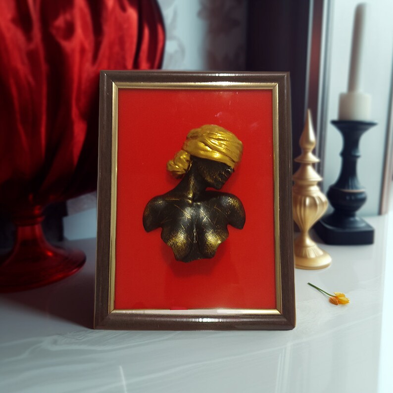May include: A framed 3D sculpture of a person's bust with a gold head covering and a gold-painted body against a red background.