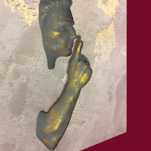 May include: A grey and gold sculpture of a person with their finger to their lips, as if to say "shhh". The sculpture is mounted on a textured grey wall.