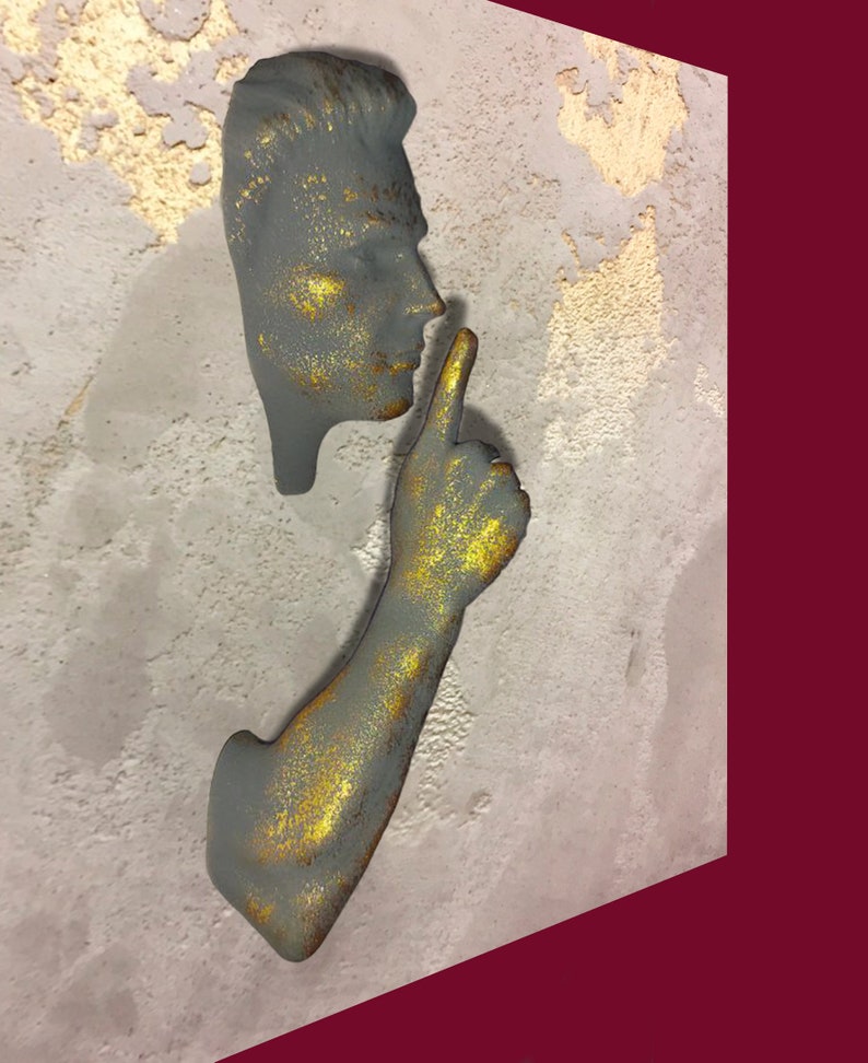 May include: A grey and gold sculpture of a person with their finger to their lips, as if to say "shhh". The sculpture is mounted on a textured grey wall.