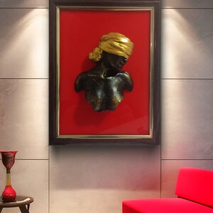 May include: A framed wall sculpture of a woman's bust with a gold head wrap. The bust is made of a dark material and is set against a red background.