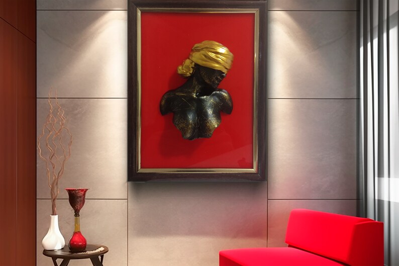 May include: A framed wall sculpture of a woman's bust with a gold head wrap. The bust is made of a dark material and is set against a red background.