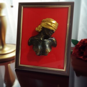 May include: A framed 3D sculpture of a person's bust with a gold head covering. The sculpture is set against a red background.