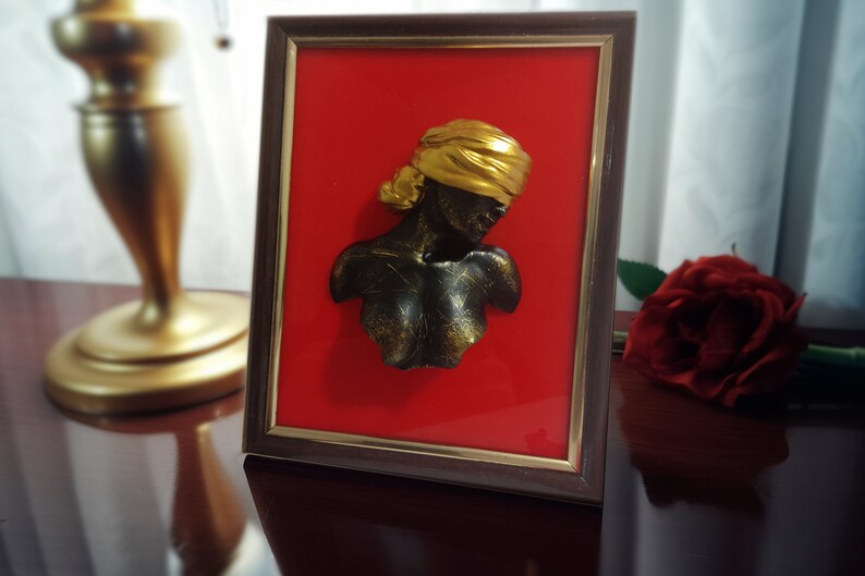 May include: A framed 3D sculpture of a person's bust with a gold head covering. The sculpture is set against a red background.
