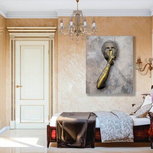 May include: A gold-toned sculpture of a person with their finger to their lips, suggesting silence. The sculpture is mounted on a textured, grey canvas.