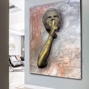 May include: A 3D wall sculpture of a person with their finger to their lips, suggesting silence. The sculpture is painted in gold and grey tones and is set against a textured background with shades of grey, brown, and pink.