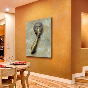 May include: A gold-toned sculpture of a person with their finger to their lips, suggesting silence. The sculpture is mounted on a textured, light blue background.