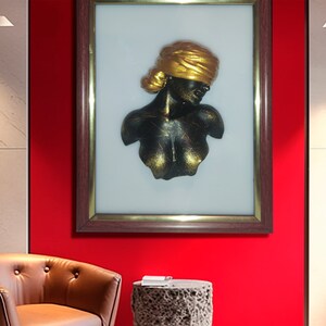 May include: A framed 3D wall sculpture of a woman's bust with a gold head wrap. The sculpture is black with gold accents and is set against a white background.