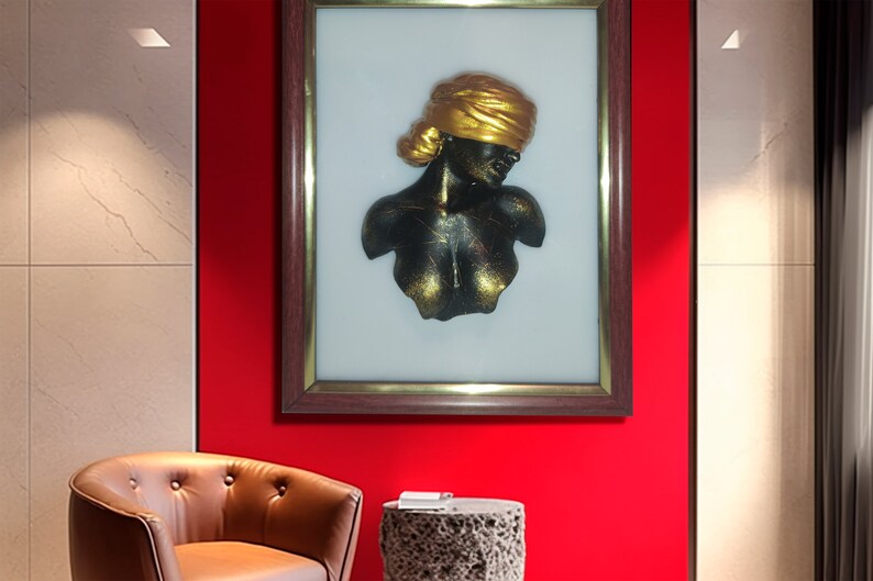 May include: A framed 3D wall sculpture of a woman's bust with a gold head wrap. The sculpture is black with gold accents and is set against a white background.