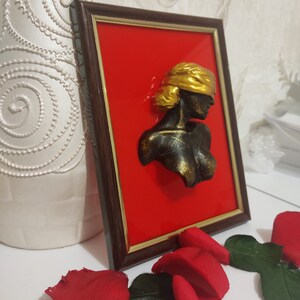 May include: A framed 3D wall sculpture of a woman's bust with a gold blindfold. The sculpture is painted black and gold and is set against a red background.