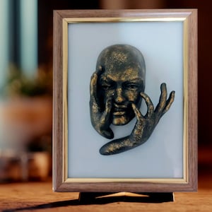 May include: A framed wall sculpture of a gold-toned face with hands covering the mouth and eyes. The sculpture is set against a white background.