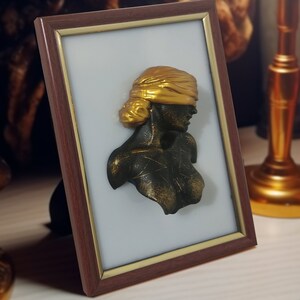 May include: A framed bronze bust of a person wearing a gold blindfold. The bust is set against a white background.